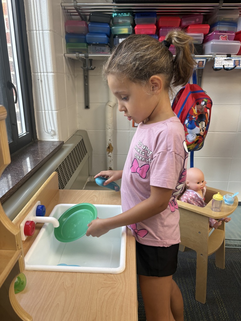 Young girl washing a green plastic plate at a small play kitchen sink in a classroom setting.