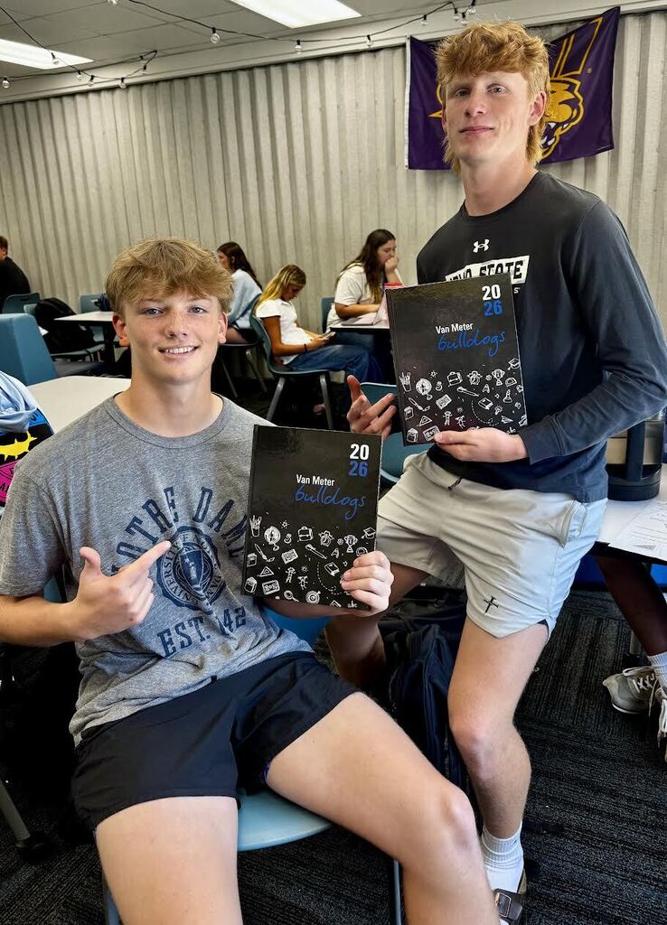 Two teenage boys in a classroom proudly hold 2026 yearbooks with 'Van Meter Bulldogs' on the cover, smiling at the camera.