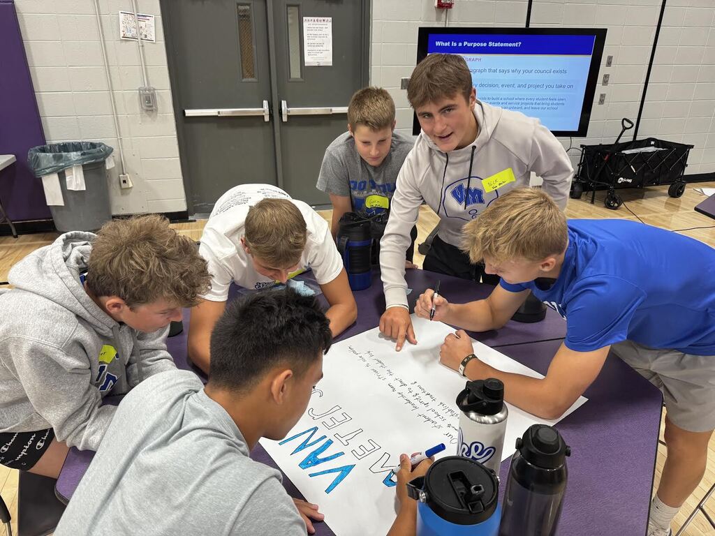 A group of six teenage boys collaborating on a large poster in a gymnasium setting, focused on writing and drawing.