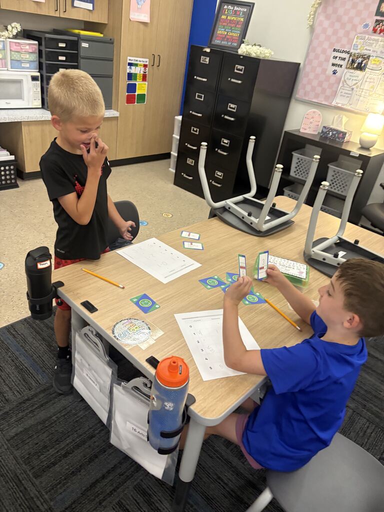Two students practice math together as one holds up number cards and the other studies a worksheet.