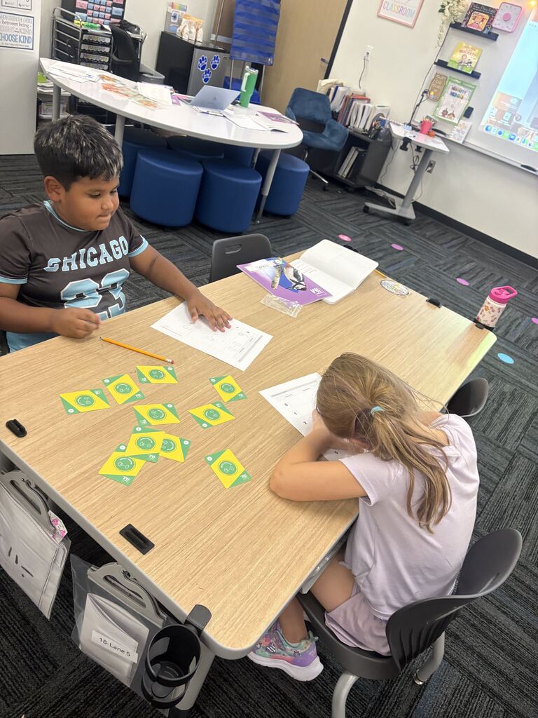 Two students complete math worksheets at a table surrounded by green and yellow learning cards.
