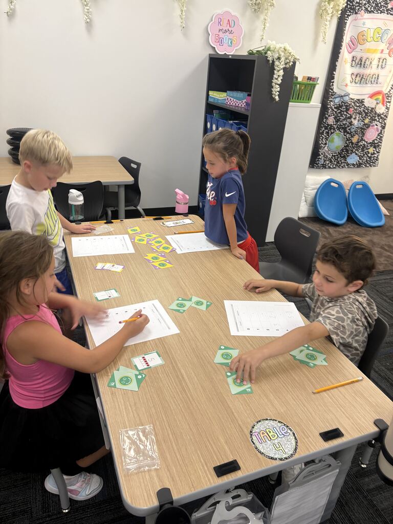Four students work around a classroom table using colorful math cards, worksheets, and pencils.