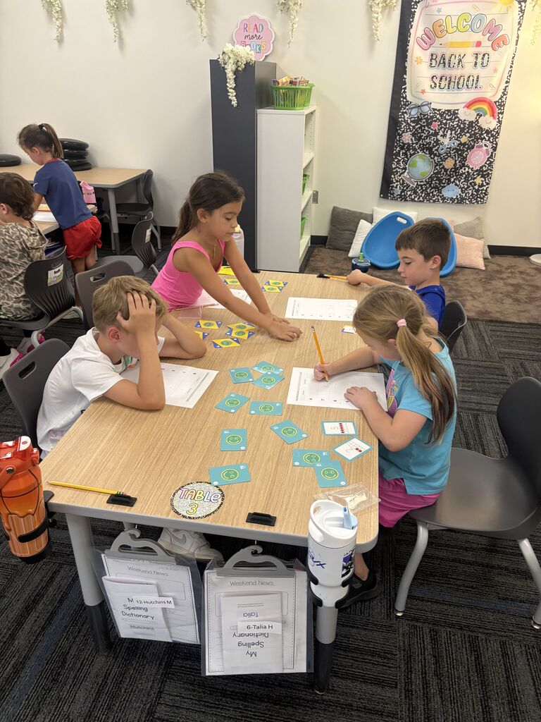 Four students practice math at a classroom table with colorful learning cards, worksheets, and pencils.