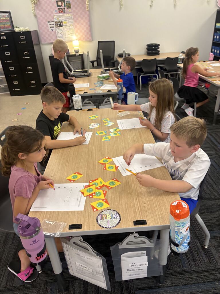 Students work in pairs at classroom tables, using math cards, worksheets, and pencils to solve problems.