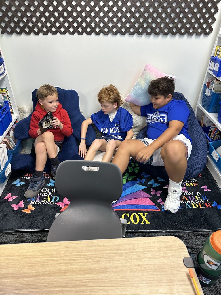 Three boys sit together in a cozy classroom reading corner, one holding a book while the others look on.