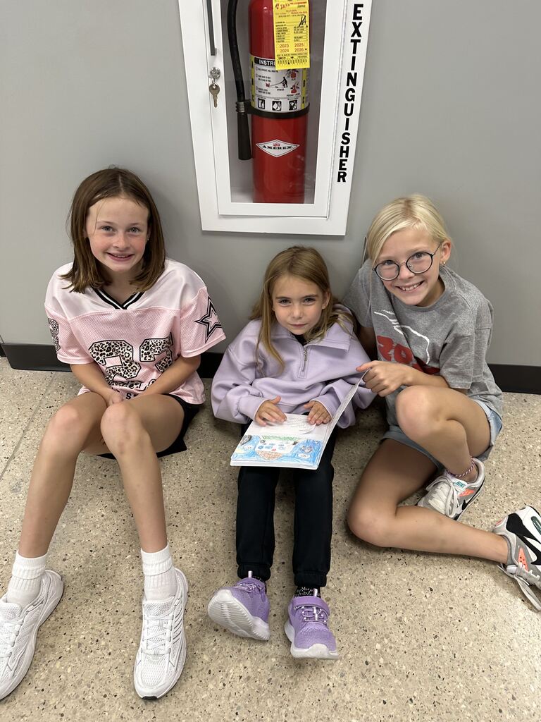 Three smiling girls sit together on a school hallway floor, with the center student holding an illustrated paper.
