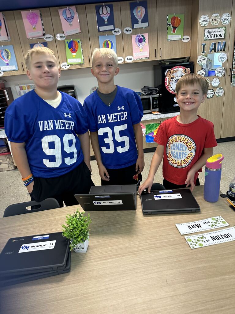 Three smiling boys stand behind a classroom table with Chromebooks, surrounded by colorful student artwork.