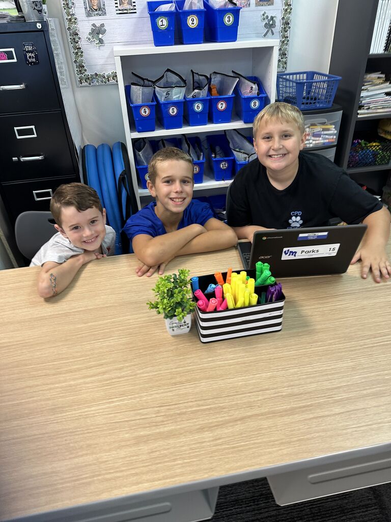 Three smiling boys gather around a classroom table with a Chromebook and colorful markers.