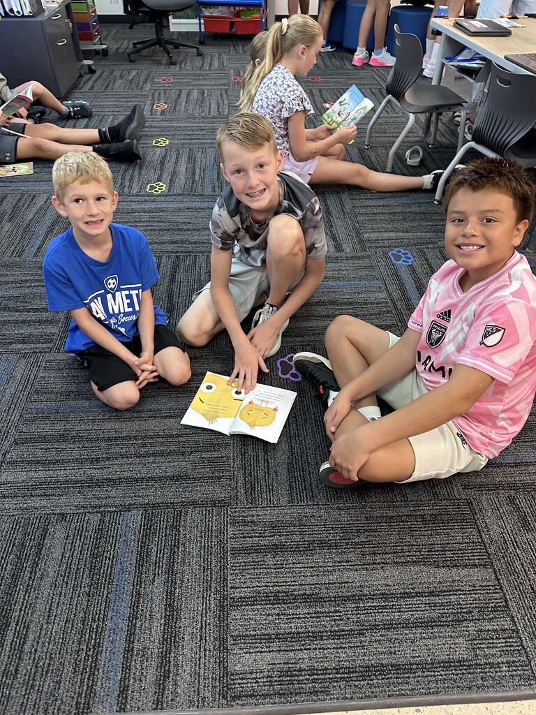 Three smiling boys sit on a classroom floor with an open picture book while another student reads nearby.