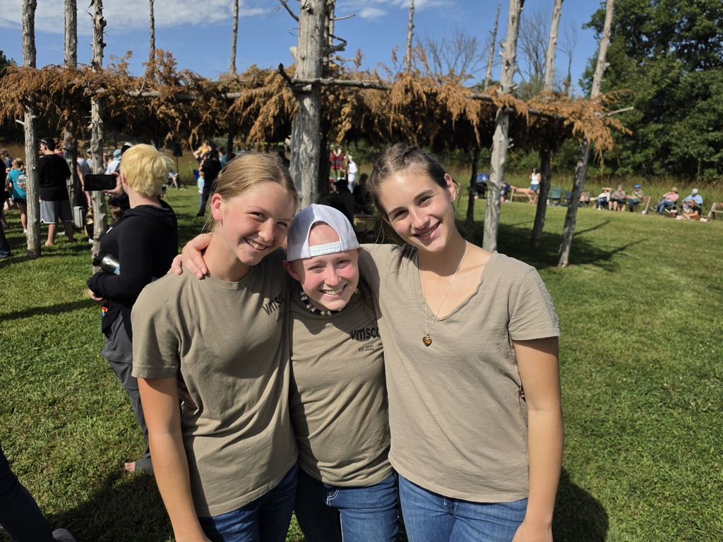 Three teenage girls smiling and posing together outdoors on a sunny day with trees and people in the background.