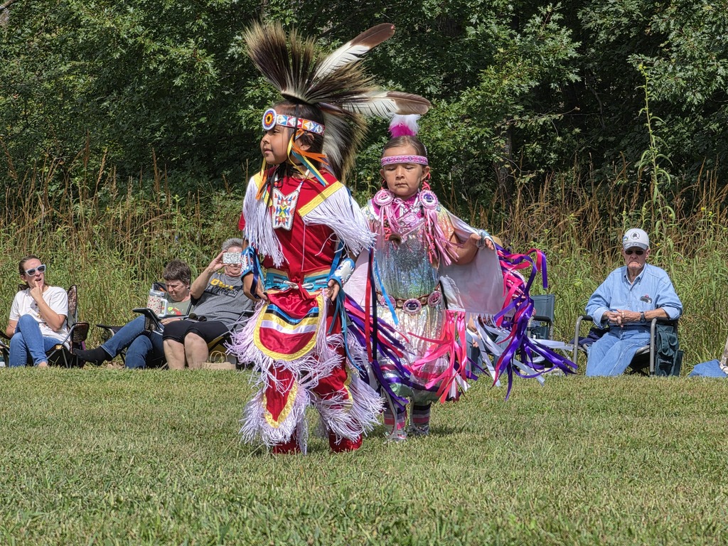 Two young Native American dancers in colorful traditional regalia perform outdoors with spectators seated behind them.