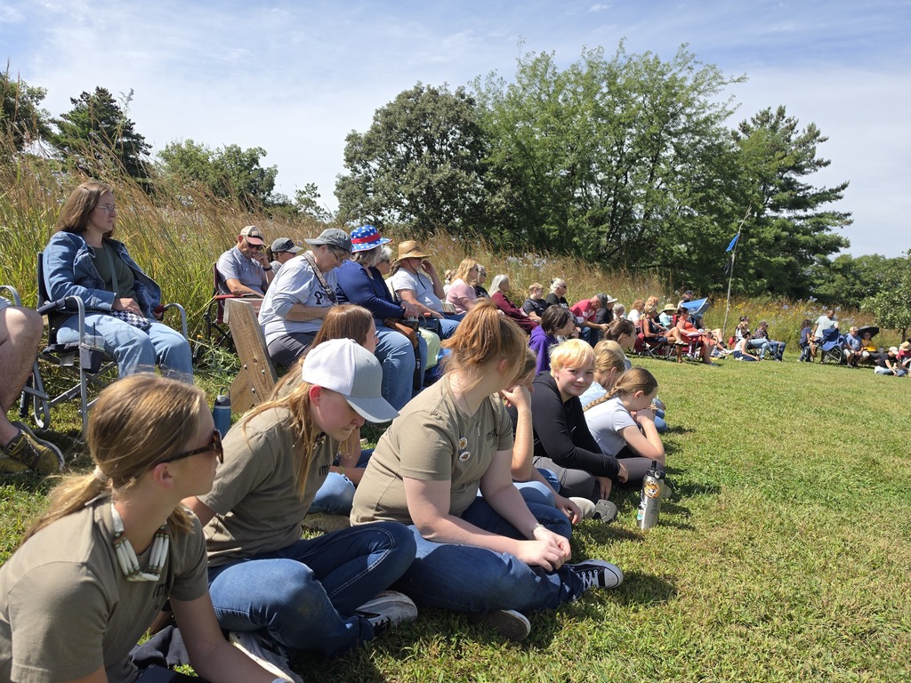 Group of people sitting on grass and folding chairs on a sunny day, watching an outdoor event in a park-like setting.