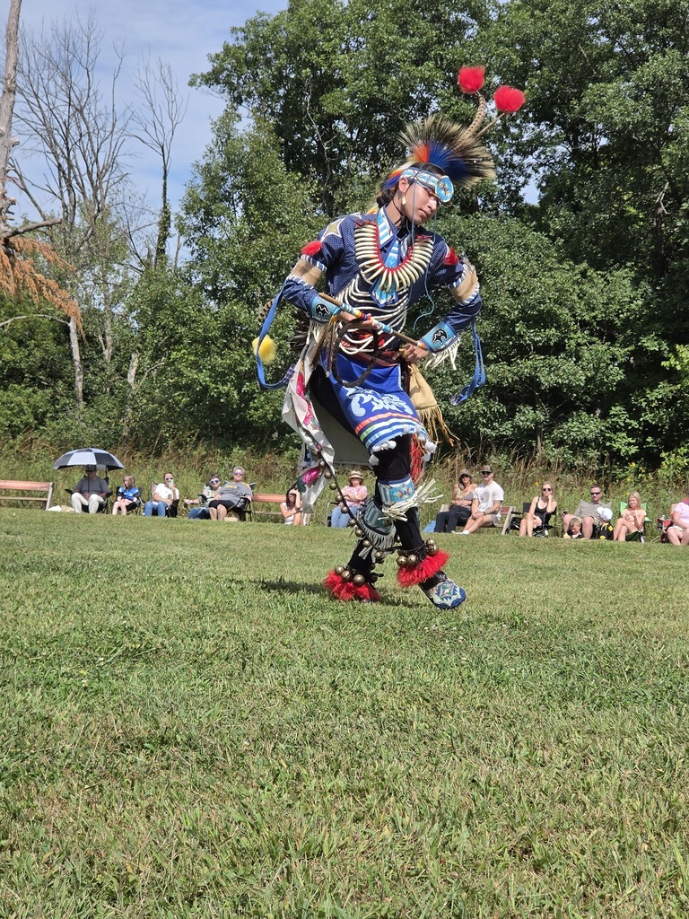 Native American dancer in vibrant traditional regalia performing outdoors on grass with spectators seated in the background.