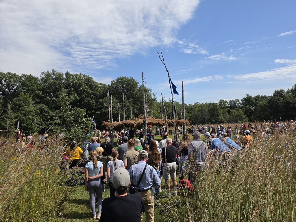 Crowd gathered outdoors near a rustic wooden structure with flags under a partly cloudy blue sky.