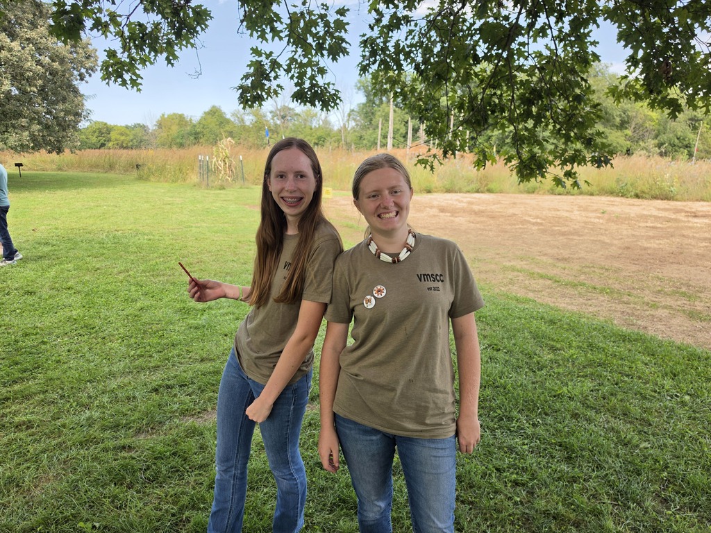 Two teenage girls smiling and standing back-to-back under a tree in a grassy park on a sunny day.