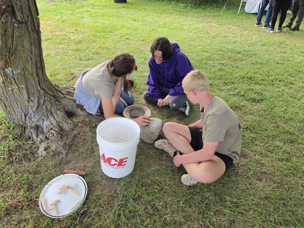 Three young people sitting on grass near a tree, examining a round stone and a bucket labeled ACE.