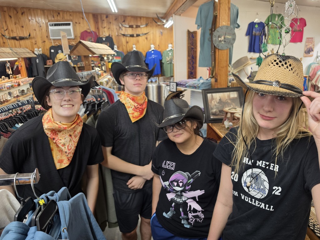 Four young people wearing cowboy hats and bandanas inside a rustic souvenir shop with wooden walls and clothing racks.