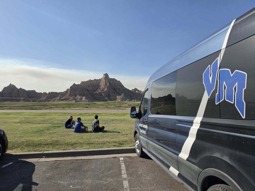 Three people sitting on grass facing rugged rock formations under a clear blue sky near a black van with blue and white lettering.
