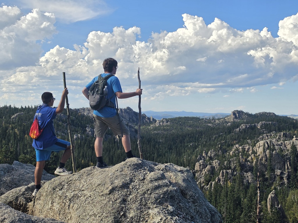 Two hikers stand on a rocky outcrop overlooking a vast forested mountain landscape under a partly cloudy sky.