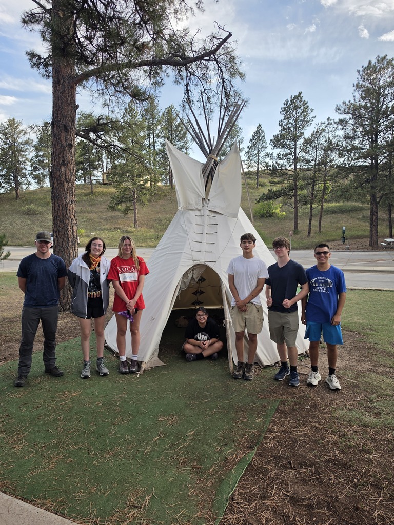 Group of seven young people posing in front of a white teepee set in a grassy, wooded area on a sunny day.