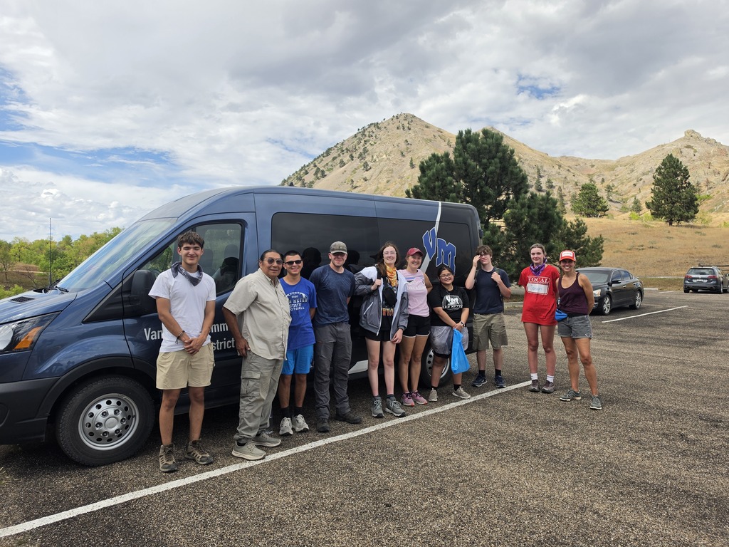 Group of ten people standing beside a dark van in a parking lot with mountains and trees in the background under a cloudy sky.