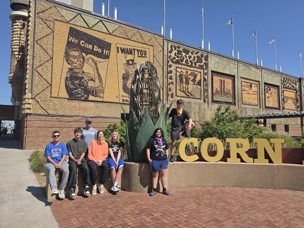 Group of seven people posing in front of a large corn sculpture and murals depicting historical agricultural scenes on a sunny day.