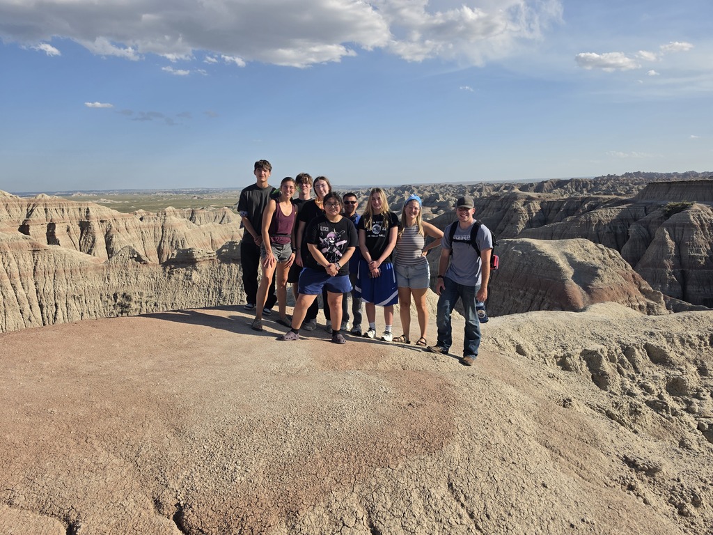 Group of nine hikers posing on a rocky plateau with expansive rugged canyon landscape under a partly cloudy sky.