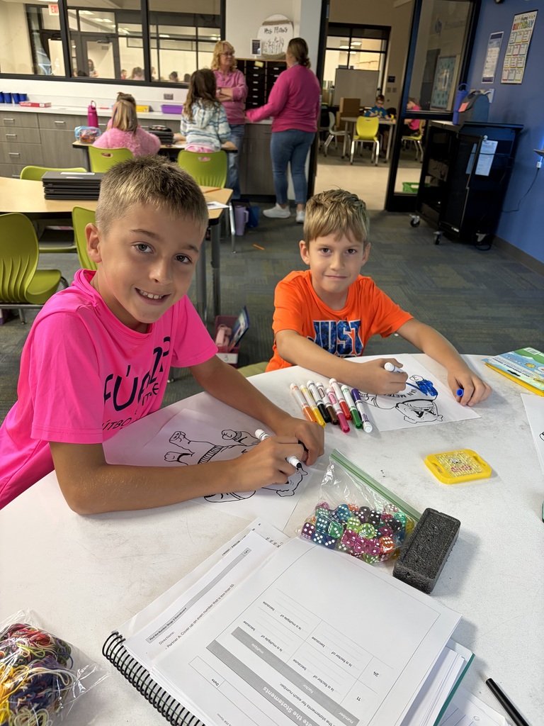 Two boys coloring superhero drawings at a classroom table with markers and school supplies nearby.