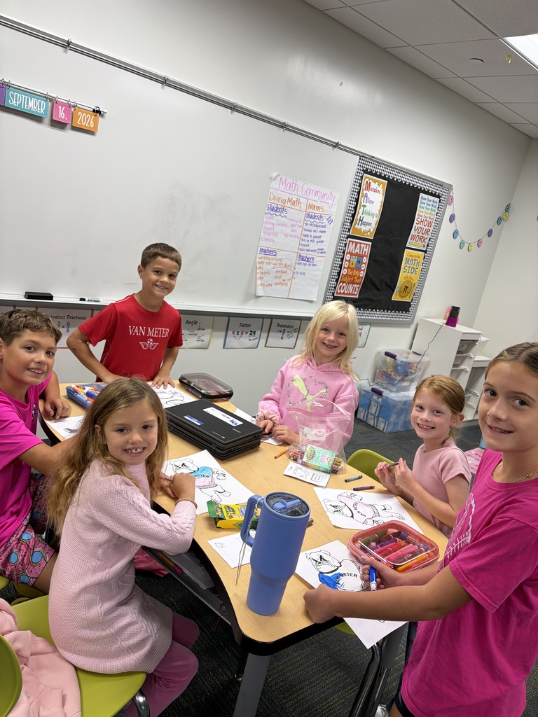 Six young students smiling and coloring at a classroom table filled with art supplies and papers.