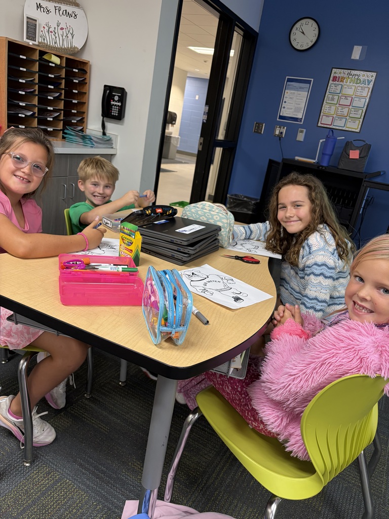 Four students sitting around a classroom table engaged in coloring and drawing activities with art supplies scattered on the table.