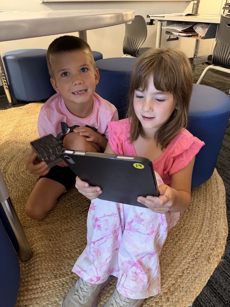 Two children sit together on a woven classroom rug, smiling and looking at a tablet held between them.