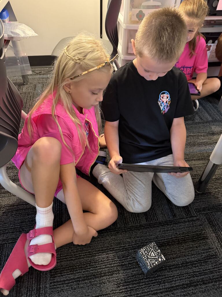 Two children sit side by side on the classroom floor, focused on a tablet with a patterned cube nearby.