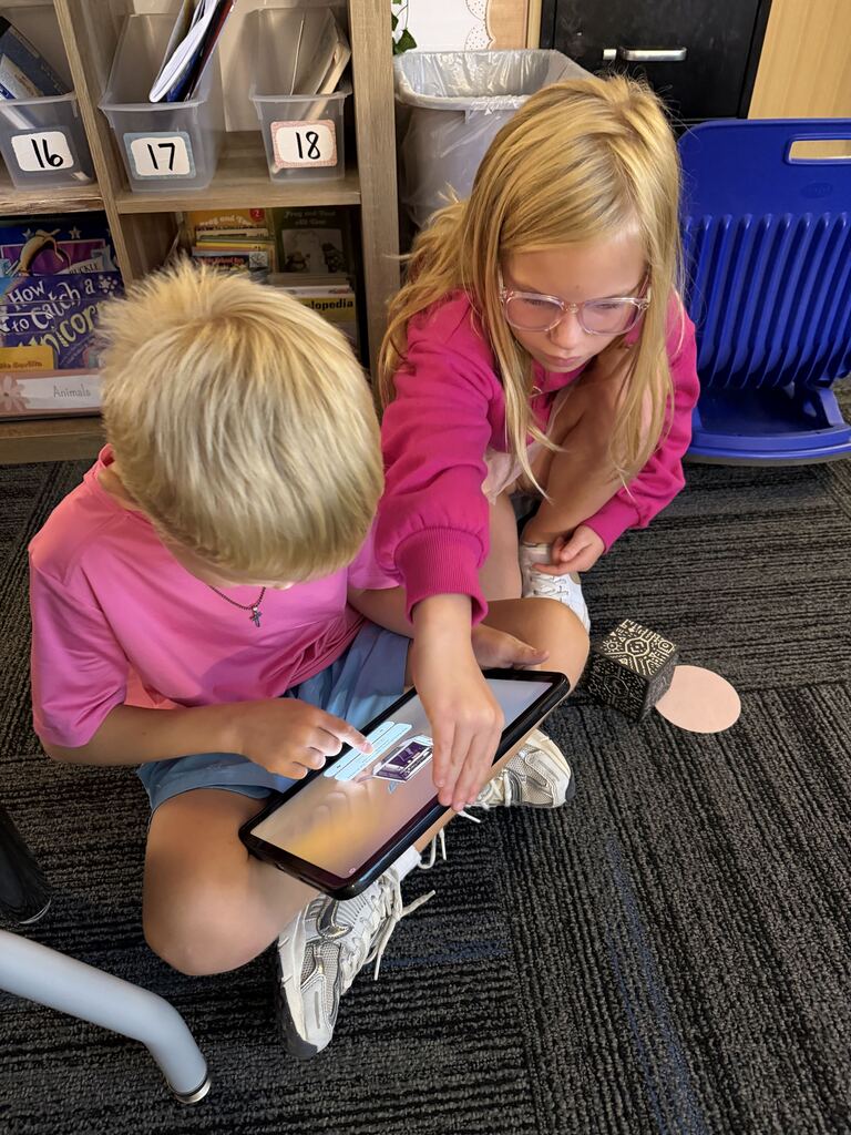 Two children in pink sit on the classroom floor, tapping a tablet together beside a patterned activity cube.