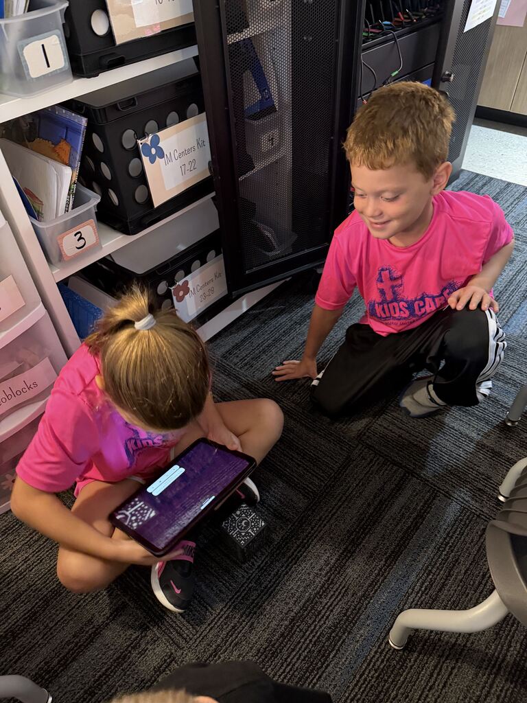 Two children in bright pink shirts sit on the classroom floor, working with a tablet and patterned cube.