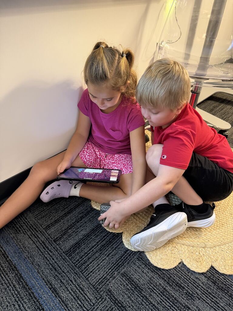 Two children sit closely on a classroom rug, looking at a tablet while one reaches toward a patterned cube.