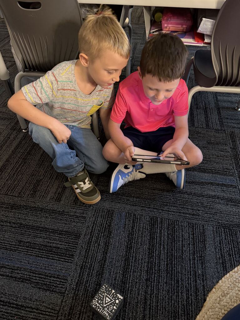 Two children sit close together on the classroom floor, concentrating on a tablet with a patterned cube in front of them.