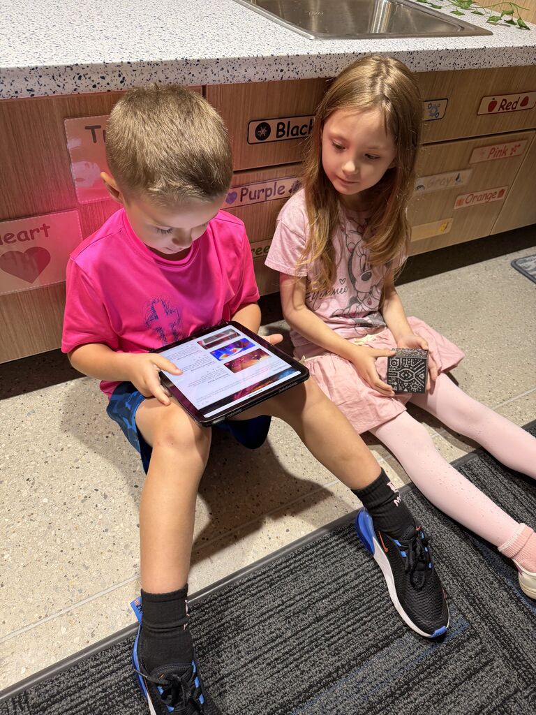 Two children sit by classroom cabinets, using a tablet and patterned activity cube together.