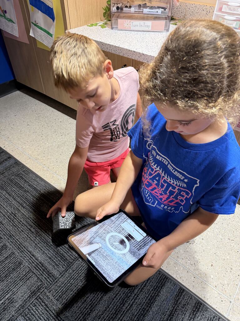 Two children kneel together in the classroom, using a tablet and patterned activity cube.