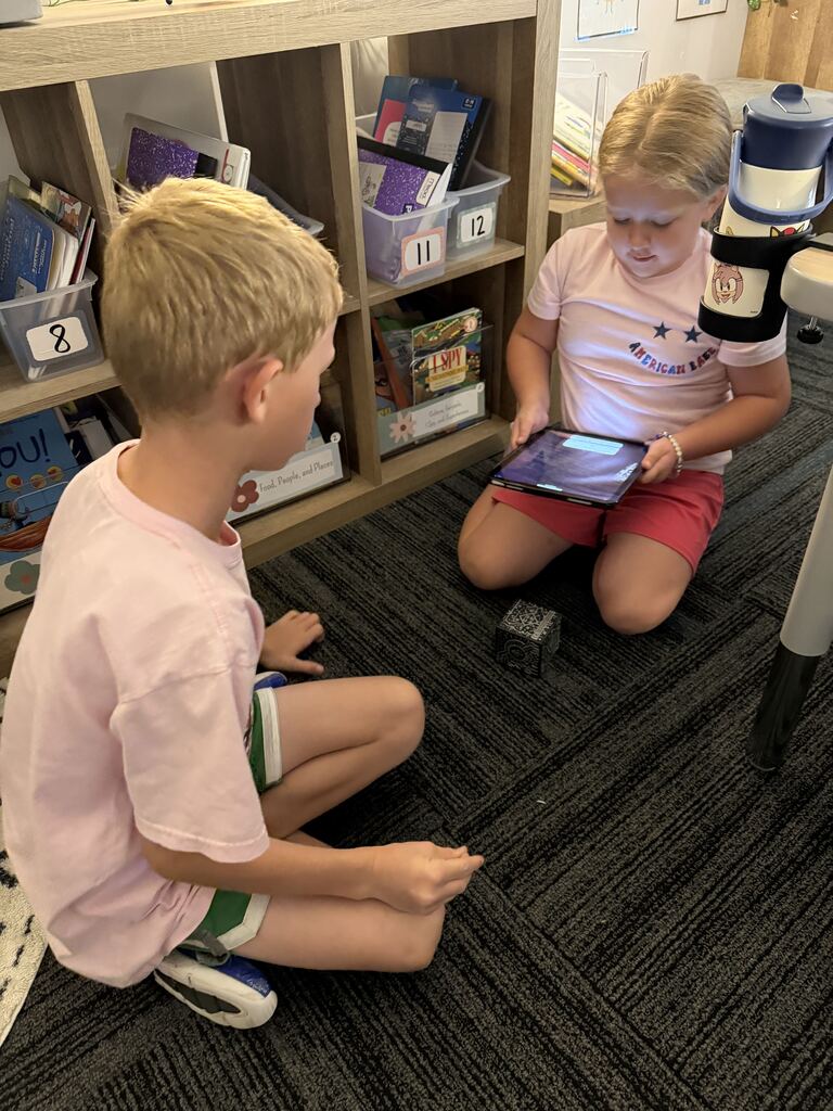 Two children sit facing each other near a classroom bookshelf, using a tablet and patterned activity cube.