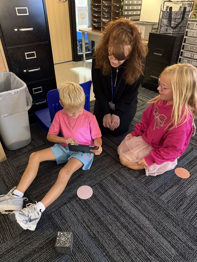 A teacher kneels beside two children in pink as they gather around a tablet during a classroom activity.