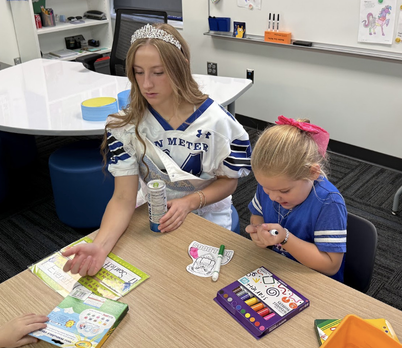 An older student wearing a tiara helps a young child complete a coloring activity with markers at a classroom table.