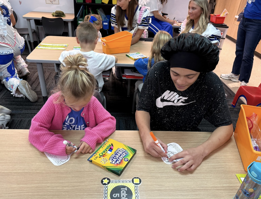 A young child and an older student color paper cutouts with markers while other students work together in the classroom.
