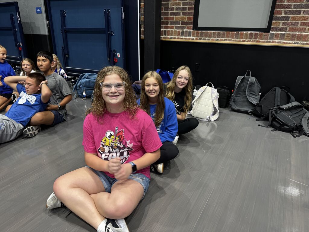 Three smiling students sit together on a gym floor while other students gather nearby with backpacks along the wall.