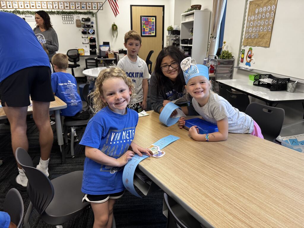Smiling elementary students and older student helpers gather around a classroom table while making blue paper crafts.