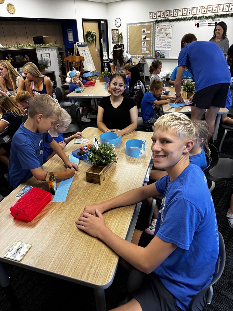 Students and older helpers work together on crafts at classroom tables, with two smiling participants facing the camera.