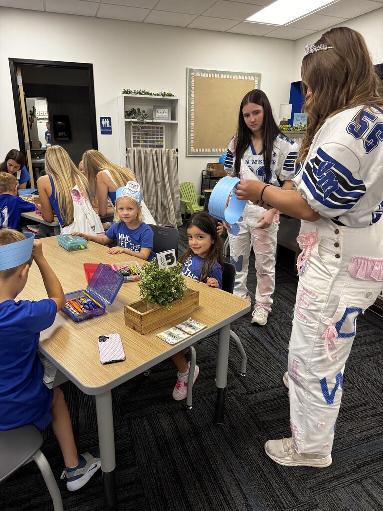 Older student helpers assist young children with blue paper crafts and coloring activities around a classroom table.