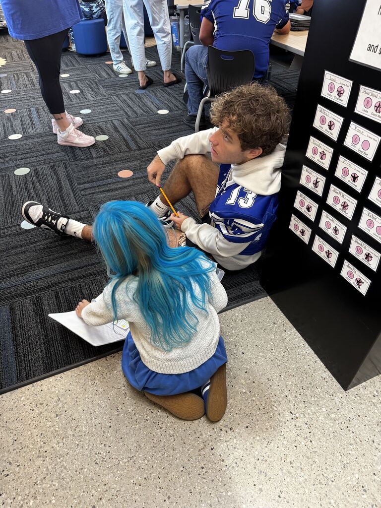 An older student sits on the classroom floor beside a child with bright blue hair as they work together on an activity.