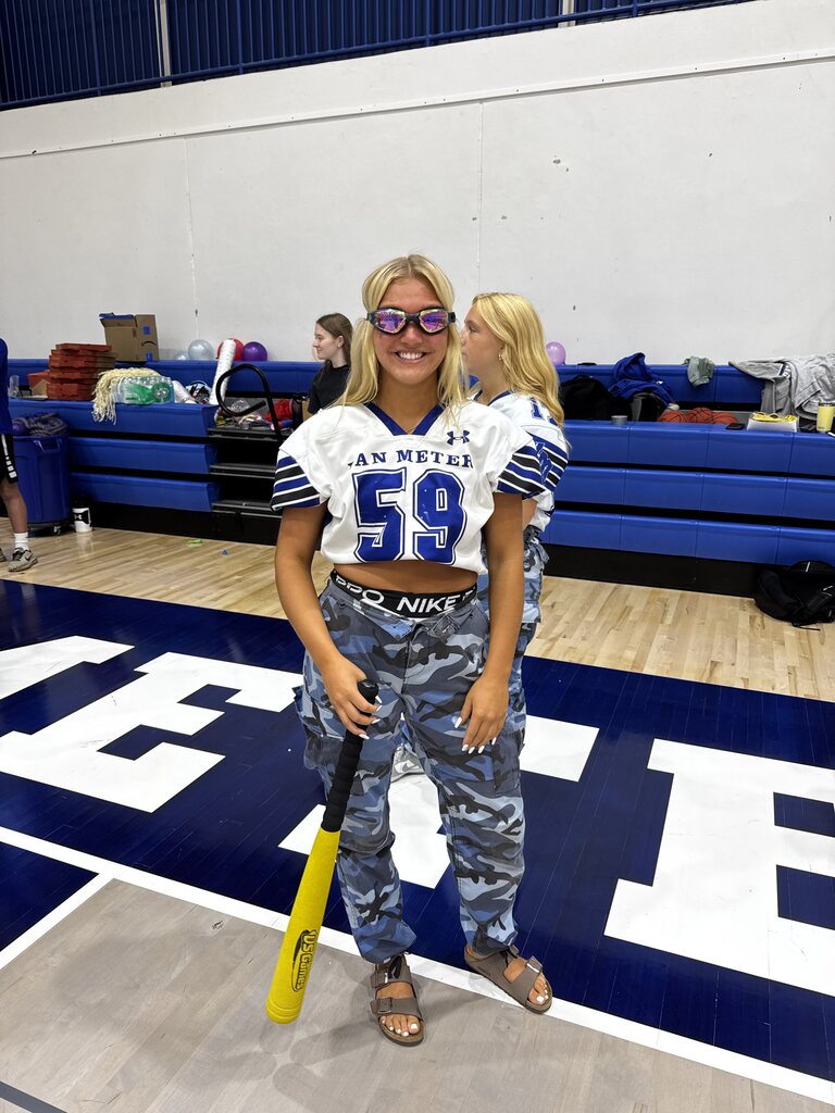 Smiling student in a white Van Meter jersey and blue camouflage pants poses with a yellow plastic bat.