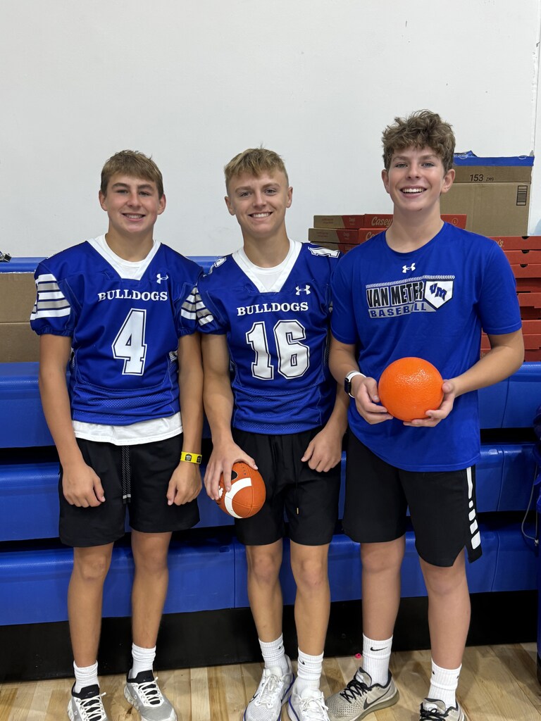 Three students pose by the gym bleachers in Bulldogs and Van Meter athletic gear, holding a football and orange ball.