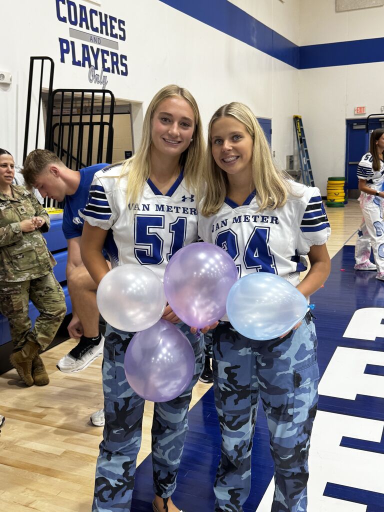 Two students in white Van Meter football jerseys and blue camouflage pants pose with pastel balloons in the gym.